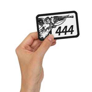 May include: A rectangular patch with a black border and a white background. The patch features a black illustration of an angel playing a trumpet, with the number "444" in bold black font. The patch is held in a hand.