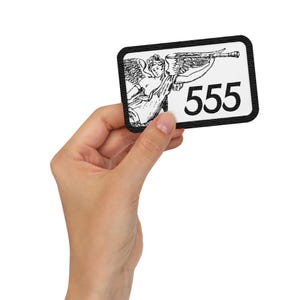 Può includere: Una toppa rettangolare con bordo nero. Presenta un'illustrazione in bianco e nero di un angelo che suona una tromba e il numero "555" in nero su sfondo bianco. La toppa è tenuta da una mano.