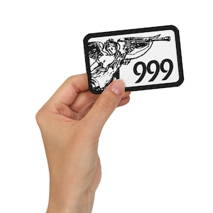 Puede incluir: Un parche rectangular con borde negro, con una ilustración en blanco y negro de un ángel tocando una trompeta, con el número "999" en negrita negra. El parche se sostiene en una mano.