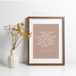 May include: A framed print with the quote "MEN ARE MORE INTERESTING IN BOOKS THAN THEY ARE IN REAL LIFE." The print has a brown frame and a light brown background. A white vase with yellow flowers sits to the left of the print.