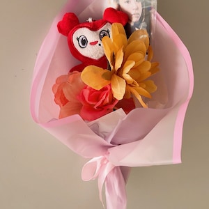 Puede incluir: Un ramo rosa con un juguete de peluche rojo, flores amarillas y rojas y una foto de una persona dentro de una funda de pl&aacute;stico transparente.