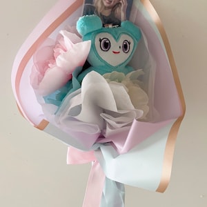 Puede incluir: Un ramo de flores en tonos pastel azul, blanco y rosa con un peluche azul claro con forma de coraz&oacute;n y ojos grandes. Una tarjeta de foto con una mujer est&aacute; metida en el ramo.
