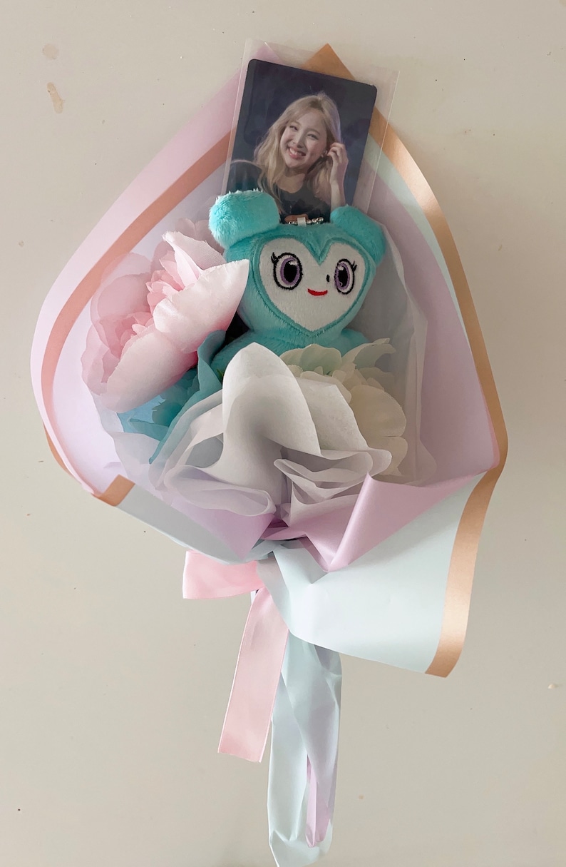 Puede incluir: Un ramo de flores en tonos pastel azul, blanco y rosa con un peluche azul claro con forma de coraz&oacute;n y ojos grandes. Una tarjeta de foto con una mujer est&aacute; metida en el ramo.