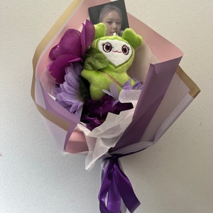 Puede incluir: Un ramo de flores moradas envuelto en papel rosa y morado con un juguete de peluche verde y una tarjeta con foto.