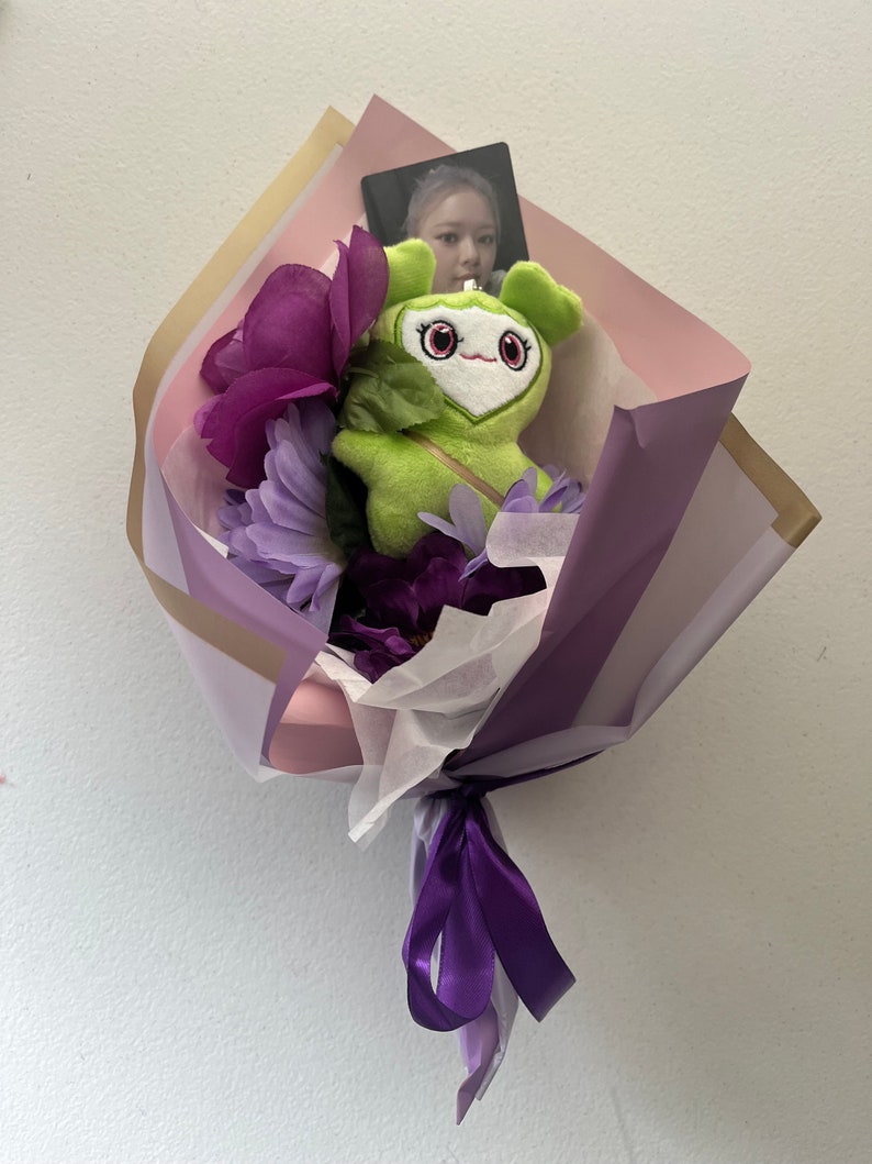 Puede incluir: Un ramo de flores moradas envuelto en papel rosa y morado con un juguete de peluche verde y una tarjeta con foto.