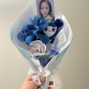 Puede incluir: Un ramo de flores artificiales azul y blanco con un juguete de peluche azul y una tarjeta de foto de una mujer. El ramo est&aacute; envuelto en papel de seda blanco y atado con una cinta blanca.
