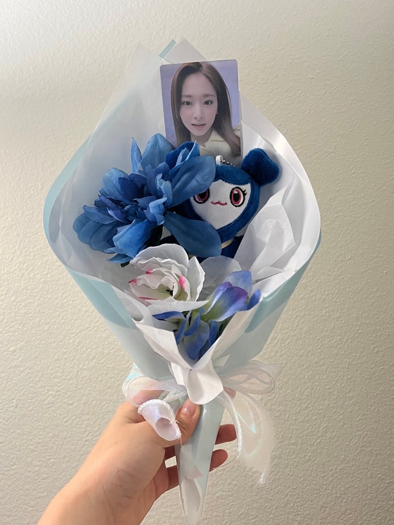Puede incluir: Un ramo de flores artificiales azul y blanco con un juguete de peluche azul y una tarjeta de foto de una mujer. El ramo est&aacute; envuelto en papel de seda blanco y atado con una cinta blanca.