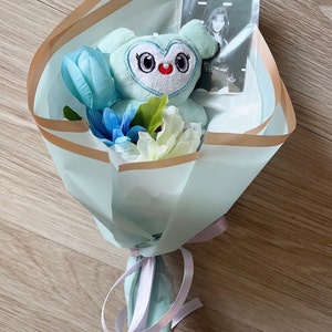 Puede incluir: Un juguete de peluche azul claro con forma de b&uacute;ho con ojos grandes y un coraz&oacute;n rojo en el pecho est&aacute; en un ramo de papel azul claro y dorado con flores azules y blancas. Una tarjeta de foto con una mujer con una camisa blanca tambi&eacute;n est&aacute; en el ramo.