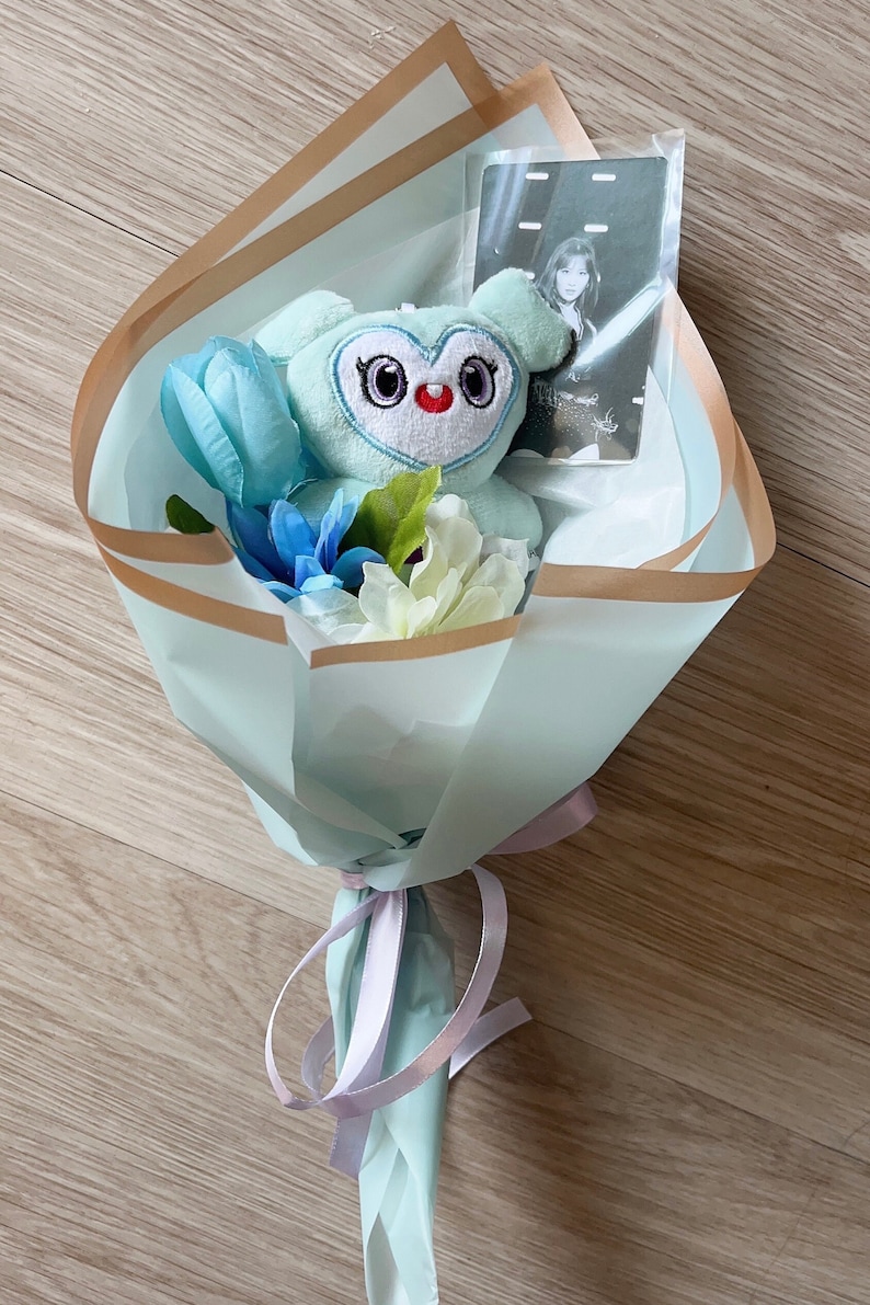 Puede incluir: Un juguete de peluche azul claro con forma de b&uacute;ho con ojos grandes y un coraz&oacute;n rojo en el pecho est&aacute; en un ramo de papel azul claro y dorado con flores azules y blancas. Una tarjeta de foto con una mujer con una camisa blanca tambi&eacute;n est&aacute; en el ramo.