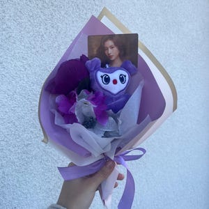 Puede incluir: Un ramo de flores morado y blanco con un juguete de peluche morado con forma de animal de dibujos animados. El juguete tiene ojos grandes y una nariz roja. El ramo est&aacute; atado con una cinta morada.