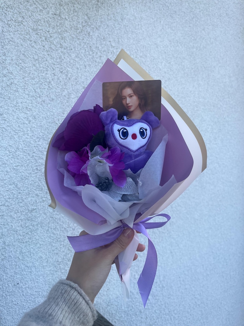 Puede incluir: Un ramo de flores morado y blanco con un juguete de peluche morado con forma de animal de dibujos animados. El juguete tiene ojos grandes y una nariz roja. El ramo est&aacute; atado con una cinta morada.