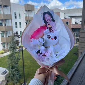 Puede incluir: Un ramo blanco con flores rosas y blancas, un juguete de peluche blanco con un coraz&oacute;n en el pecho y una tarjeta de foto de una mujer sosteniendo una bebida. El ramo est&aacute; atado con una cinta marr&oacute;n.