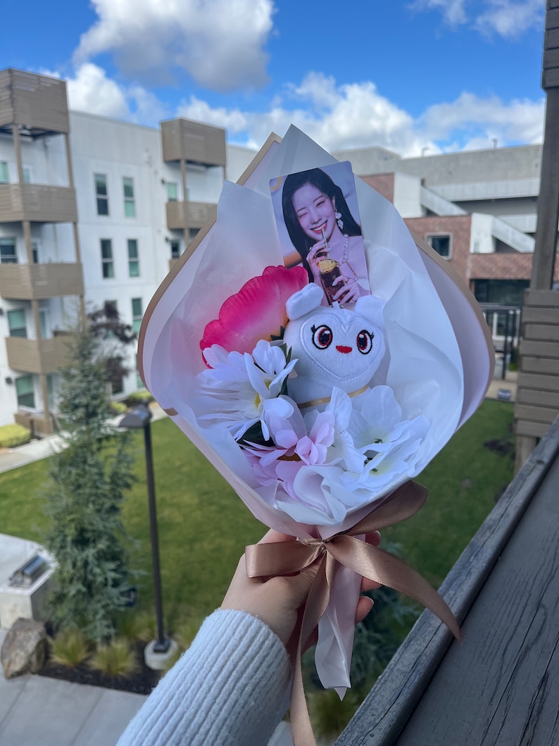 Puede incluir: Un ramo blanco con flores rosas y blancas, un juguete de peluche blanco con un coraz&oacute;n en el pecho y una tarjeta de foto de una mujer sosteniendo una bebida. El ramo est&aacute; atado con una cinta marr&oacute;n.