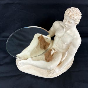 May include: A cream-coloured sculpture of a reclining figure holding a round mirror. The figure has curly hair and is resting on a textured base. A few brown leaves are placed on the sculpture. The background is a solid black.