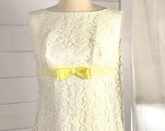 May include: Ivory lace dress with yellow ribbon accents and bows. The sleeveless dress is displayed on a mannequin. The dress features a boat neckline and a straight silhouette. The yellow ribbon is placed around the waist and at the hemline.