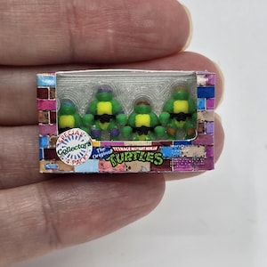 Può includere: Un set di scatole in miniatura di action figure delle Tartarughe Ninja. La scatola presenta le parole "Special Collectors Pack" e "The Original Turtles". Le figure sono verdi con maschere colorate.
