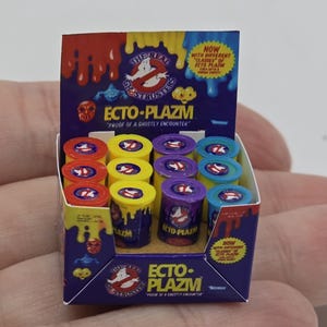 May include: Miniature display box of Ecto-Plazm, a Ghostbusters-themed product. The box is purple, yellow, and red, with the Ghostbusters logo and text. The display contains several small, colorful containers of Ecto-Plazm.