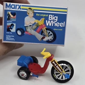 Puede incluir: Un modelo en miniatura del juguete Big Wheel deluxe, con un marco rojo, asiento azul, manillar amarillo y ruedas negras. El embalaje muestra a un niño montando el juguete. La caja dice "Marx" y "Big Wheel".