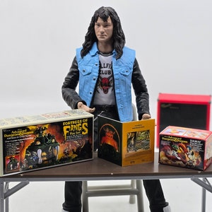 May include: A miniature figure of a person wearing a blue denim jacket with a white t-shirt that says "Hellfire Club" and a red devil graphic. The figure is sitting at a table with four game boxes. The boxes are for Dungeons and Dragons games, including "Fortress of Fangs" and "Advanced Dungeons and Dragons."