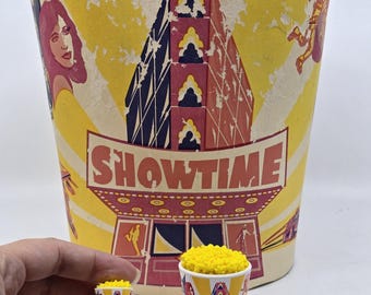 Könnte beinhalten: Ein großer Popcorn-Eimer im Vintage-Stil mit dem Wort "SHOWTIME" in roten Buchstaben. Der Eimer hat einen gelben Hintergrund mit Illustrationen einer Frau, eines Gebäudes und anderer Figuren. Zwei Miniatur-Popcorn-Eimer sind ebenfalls sichtbar.