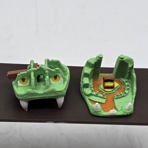 May include: A green 3D printed dragon head trap with a wooden plank and a treasure chest inside. The trap is open and the chest is visible.