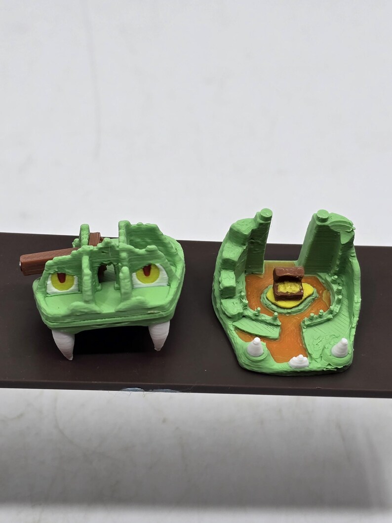 May include: A green 3D printed dragon head trap with a wooden plank and a treasure chest inside. The trap is open and the chest is visible.