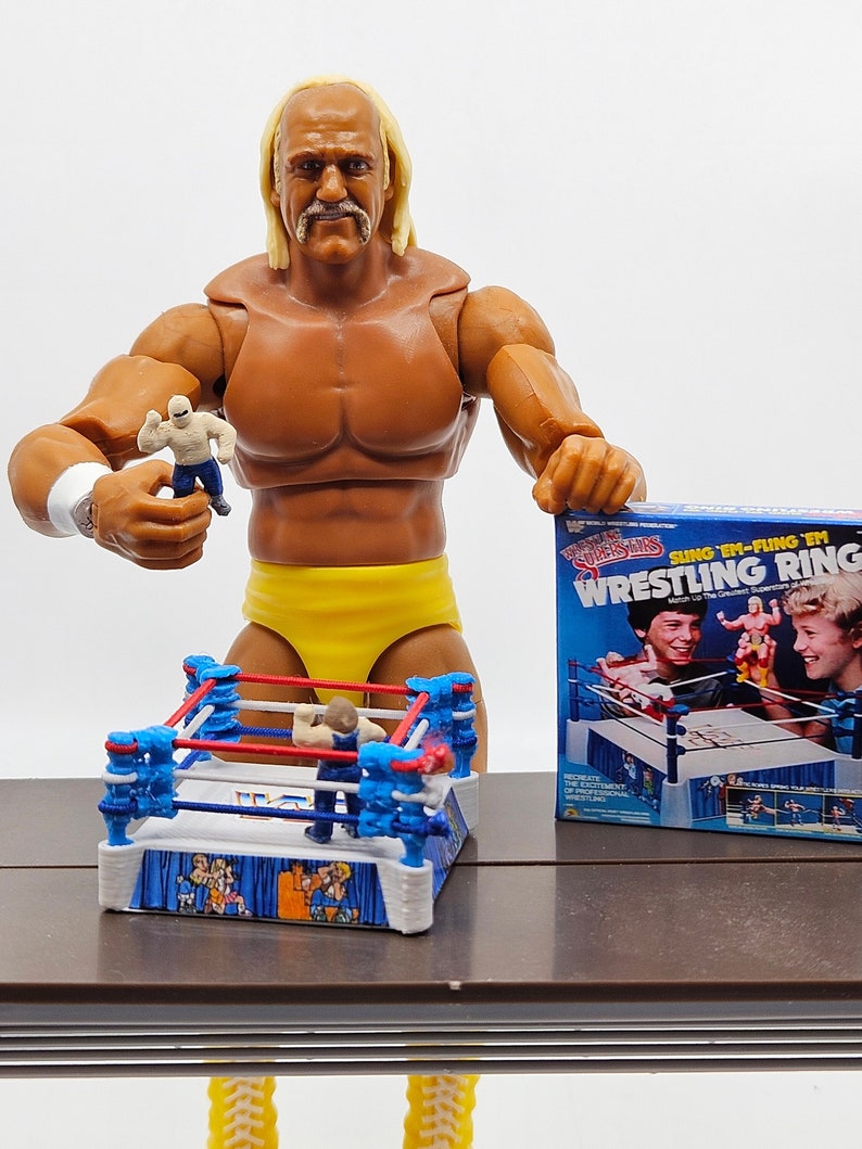 May include: A yellow and blue wrestling ring toy with a wrestler figure inside. The ring is on a brown table next to a wrestling superstar action figure holding a smaller wrestler figure. The box for the wrestling ring toy is in the background and says "Sing 'Em-Fling 'Em Wrestling Ring".
