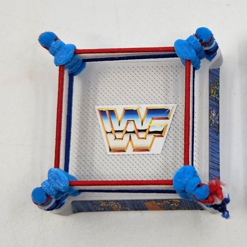 May include: A miniature wrestling ring with red, white, and blue ropes. The ring has a white background with a black and white grid pattern. The WWF logo is printed in gold on the white background.