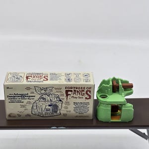 May include: A green and brown plastic model of a fortress with the text "Fortress of Fangs Play Set" on the box. The box also includes a diagram of the fortress with labeled features.