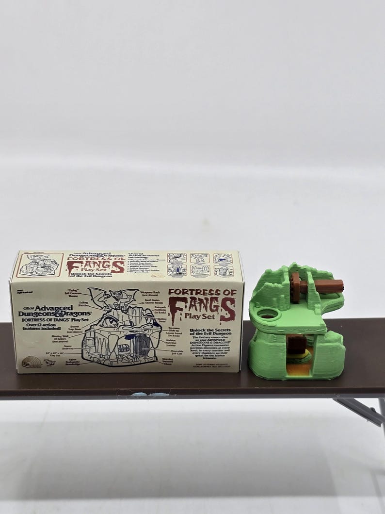 May include: A green and brown plastic model of a fortress with the text "Fortress of Fangs Play Set" on the box. The box also includes a diagram of the fortress with labeled features.
