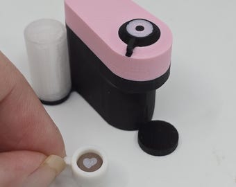 May include: A miniature coffee maker in pink and black, with a small white cup of coffee featuring a heart design. A white cylindrical object and a black disc are also present. The coffee maker is a toy or decorative item.