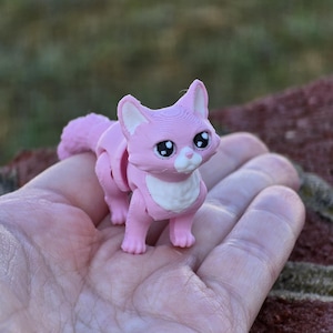 Op de afbeelding: Een roze 3D-geprint kattenbeeldje met witte accenten en grote ogen. De kat staat op zijn vier poten en heeft een lange staart.