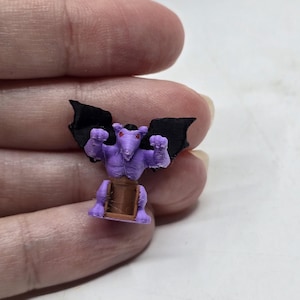 May include: A small purple gargoyle figurine with black wings and red eyes. The gargoyle is standing on a brown base.