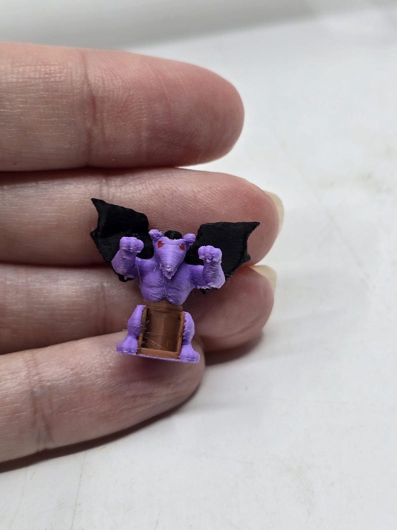 May include: A small purple gargoyle figurine with black wings and red eyes. The gargoyle is standing on a brown base.