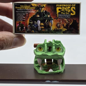 May include: A green plastic Dungeons & Dragons Fortress of Fangs playset with a dragon head design. The box art features a green dragon with fangs and a dungeon interior. The text on the box reads "Official Advanced Dungeons & Dragons Fortress of Fangs Play Set" and "Unlock the Secrets of the Evil Dungeon".