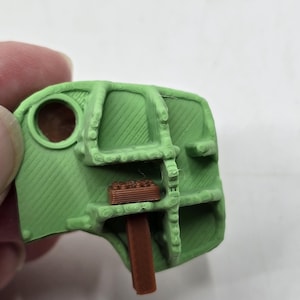 May include: A green 3D printed plastic part with a brown plastic piece inserted into it.