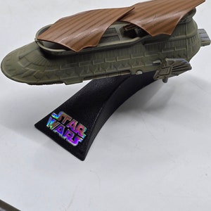 May include: A brown and grey Star Wars spaceship model with a black stand. The stand has the words "STAR WARS" printed on it.
