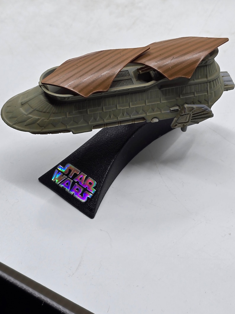 May include: A brown and grey Star Wars spaceship model with a black stand. The stand has the words "STAR WARS" printed on it.