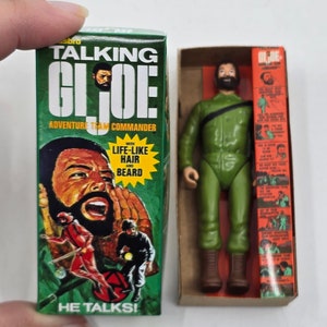 Puede incluir: Una caja verde y naranja con una figura de acci&oacute;n parlante de G.I. Joe en su interior. La caja presenta una imagen de la figura con cabello y barba realistas. El texto en la caja dice "Talking G.I. Joe Adventure Team Commander" y "He Talks!"