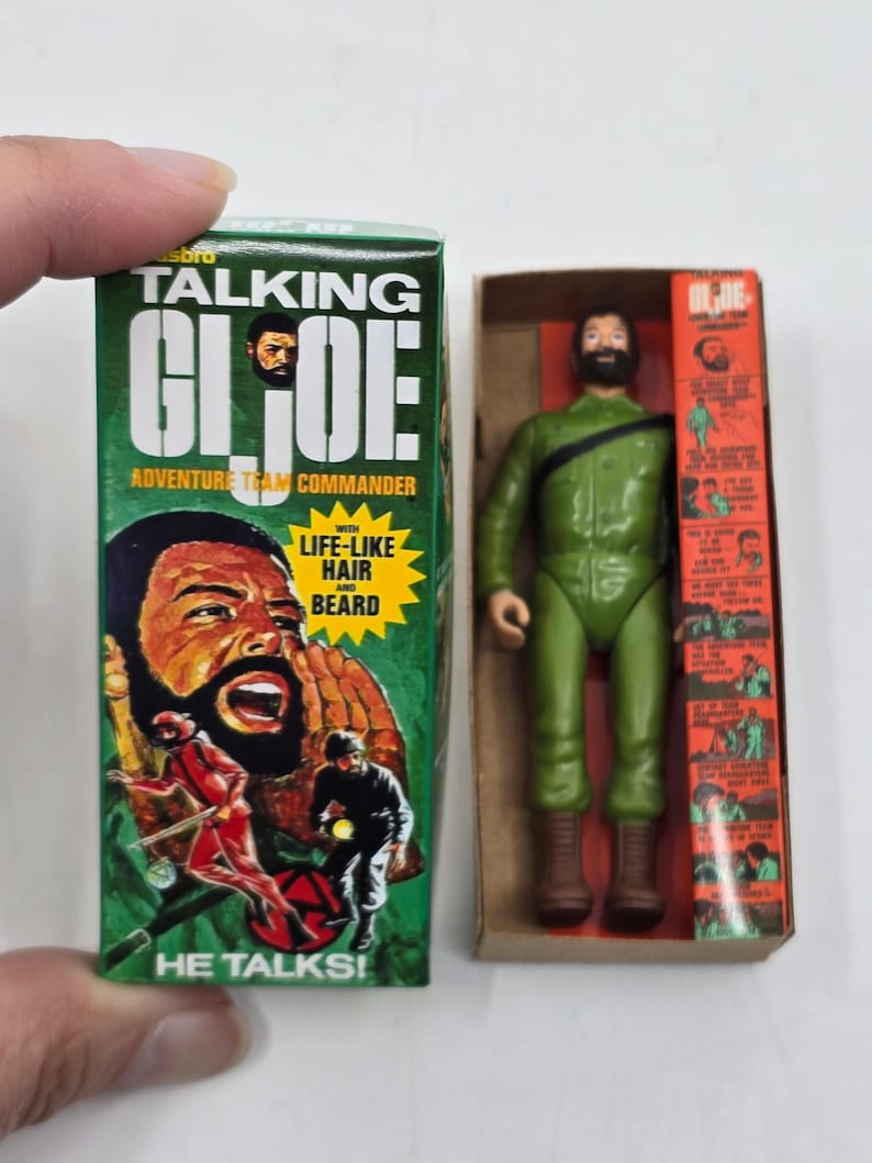 Puede incluir: Una caja verde y naranja con una figura de acci&oacute;n parlante de G.I. Joe en su interior. La caja presenta una imagen de la figura con cabello y barba realistas. El texto en la caja dice "Talking G.I. Joe Adventure Team Commander" y "He Talks!"