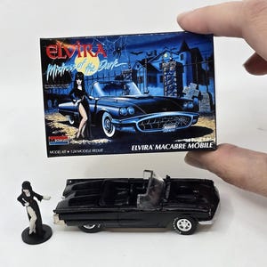 May include: A black model car kit of Elvira's car, with a small Elvira figure. The box shows a black convertible and the words "Elvira Mistress of the Dark". The model car is black with white tyres.