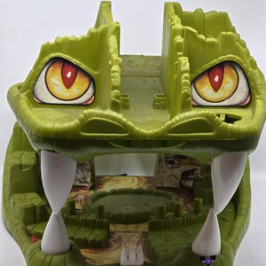 May include: Green plastic toy crocodile playset with a large open mouth, revealing a small interior play area with a red and orange lava-like floor. The crocodile has large yellow eyes with red pupils and white teeth.