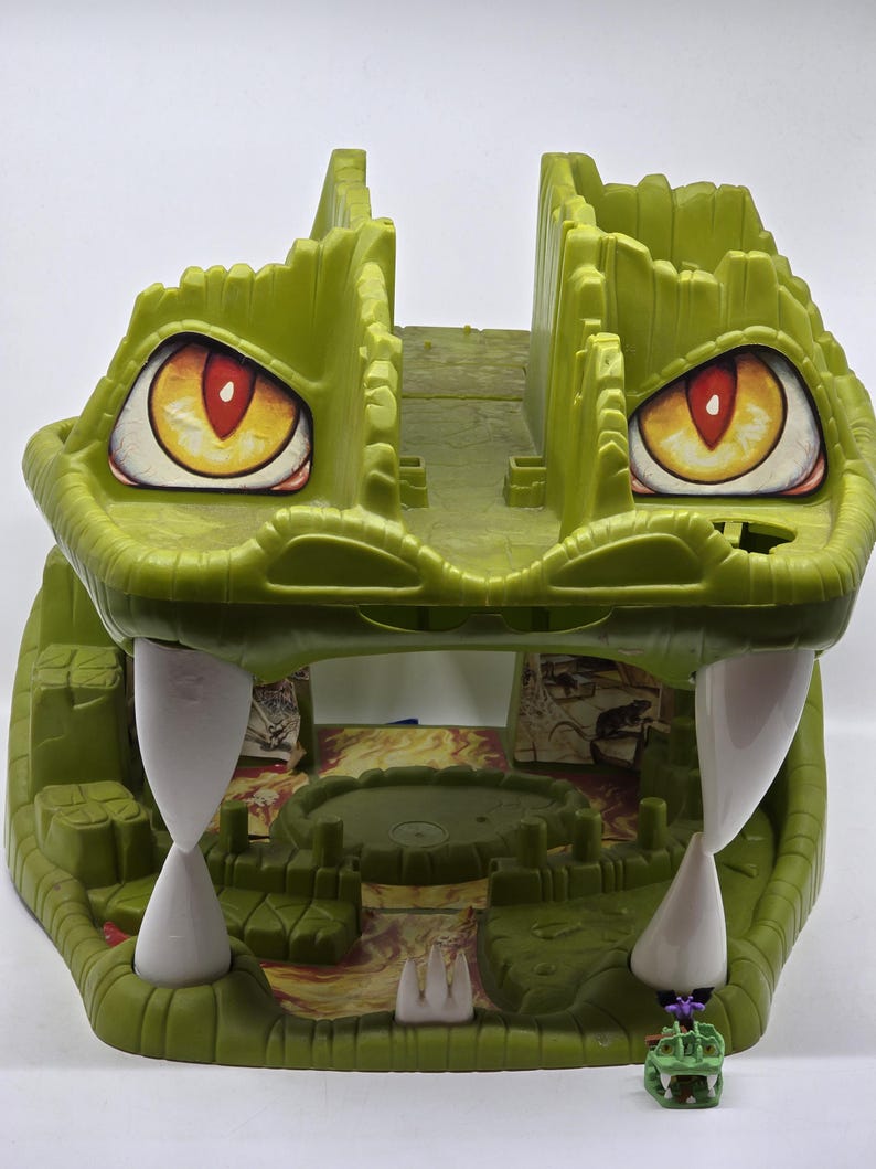 May include: Green plastic toy crocodile playset with a large open mouth, revealing a small interior play area with a red and orange lava-like floor. The crocodile has large yellow eyes with red pupils and white teeth.