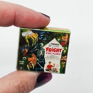 May include: A miniature "Fright Factory" toy box featuring monster illustrations. The box has a green and gold color scheme with the text "A Thingmaker Toy Featuring Fright Factory". The box is held by a person.