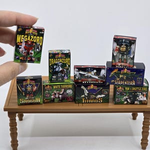 May include: Miniature toy boxes featuring various Power Rangers Zords, including Megazord, Dragonzord, and White Tigerzord. The boxes are arranged on a small wooden table, with the text "MEGAZORD", "DRAGONZORD", "WHITE TIGERZORD", "SHOGUN", "TITANUS", "SERPENTERA", and "TOR THE SHUTTLE ZORD" visible.
