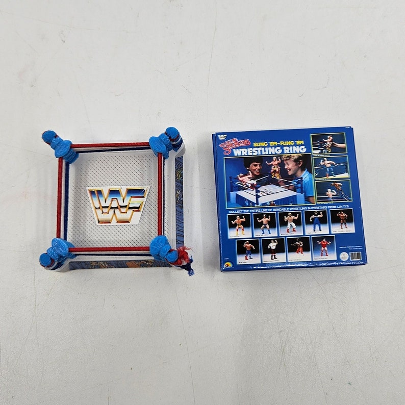 May include: A blue and red wrestling ring with a white WWF logo. The ring is part of a wrestling toy set. The box for the toy set is blue and features a picture of a wrestling ring and wrestlers. The box says "SLING 'EM-FLING 'EM WRESTLING RING".