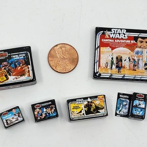 以下が含まれることがあります： スターウォーズのアクションフィギュアのおもちゃの箱のヴィンテージコレクション。箱は黒で、キャラクターと宇宙船のカラフルな画像が描かれています。箱には、「スターウォーズカンティーナアドベンチャーセット」、「スターウォーズタウタアン」、「スターウォーズダースベイダー」、「スターウォーズワムパ」、「帝国の逆襲ツインポッドクラウドカー」が含まれています。