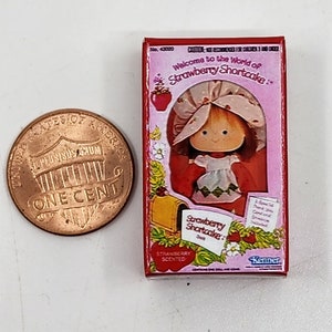 May include: A red and pink box with a picture of Strawberry Shortcake on the front. The box is labeled "Welcome to the World of Strawberry Shortcake." and "Strawberry Shortcake: Doll". The box is also labeled "Strawberry Scented".