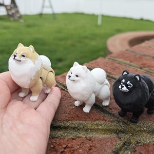 Puede incluir: Tres pequeñas figuritas de perro de peluche articuladas. Los perros están en posición de pie y mirando a la cámara. El perro de la izquierda es de color canela con pelaje blanco en el pecho y la cara. El perro del medio es blanco. El perro de la derecha es negro.