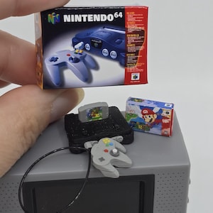 May include: A miniature Nintendo 64 console box featuring a blue console and controller graphic, alongside a small black console model with a grey controller, a black cord, and a miniature Super Mario 64 game box with a red and blue cover design.